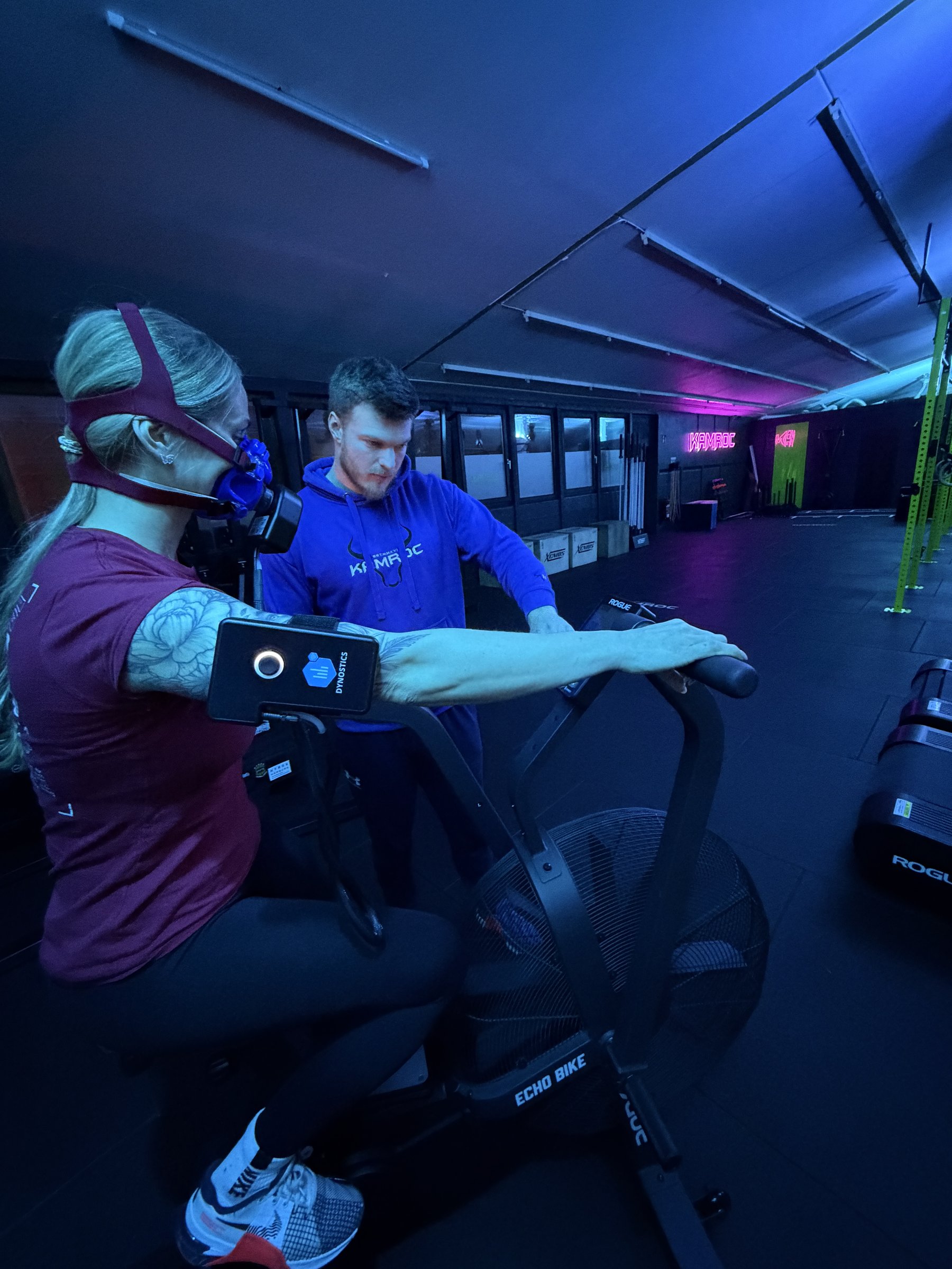 Leistungsdiagnostik bei KAMROC: Mitglied mit Atemgas-Maske auf dem Airbike, ein Coach begleitet die Messung im Studio